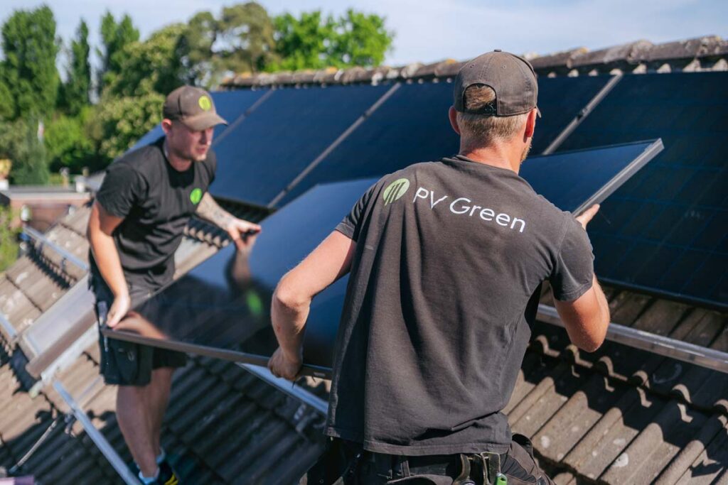 Zwei Solarteure von PV Green tragen ein PV-Modul und montieren eine PV-Anlage auf einem Dach.
